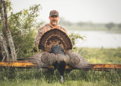 A man in camouflage clothing and a cap is holding wild turkey by their tails in an outdoor setting with grass, trees, and a body of water in the background. The man appears to be sitting on a rusty yellow metal beam while smiling at the camera.