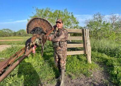 A man in camouflage hunting gear stands outdoors on a sunny day, holding a large wild turkey by a wooden fence. The background features green foliage, grass, and a clear blue sky.