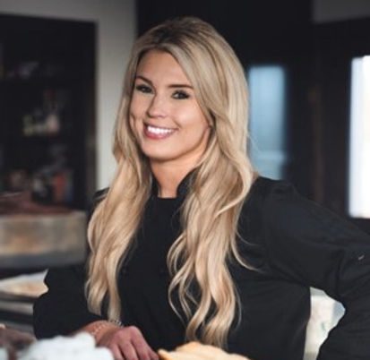 A smiling blonde woman with long hair stands in a dimly-lit room, wearing a black top. She is leaning against a counter, confidently looking at the camera. The background is slightly blurred with shelves and a bright window visible.