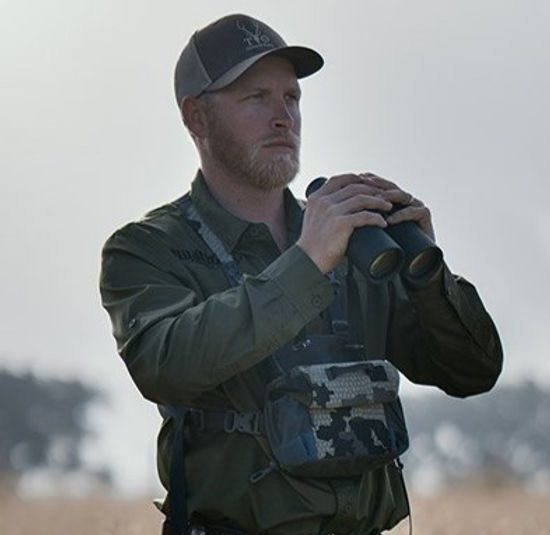 A man with a beard, wearing a cap and a green uniform, holds binoculars while looking intently into the distance. He also has a camouflage-patterned pouch strapped to his chest. The background shows a hazy outdoor setting with trees.