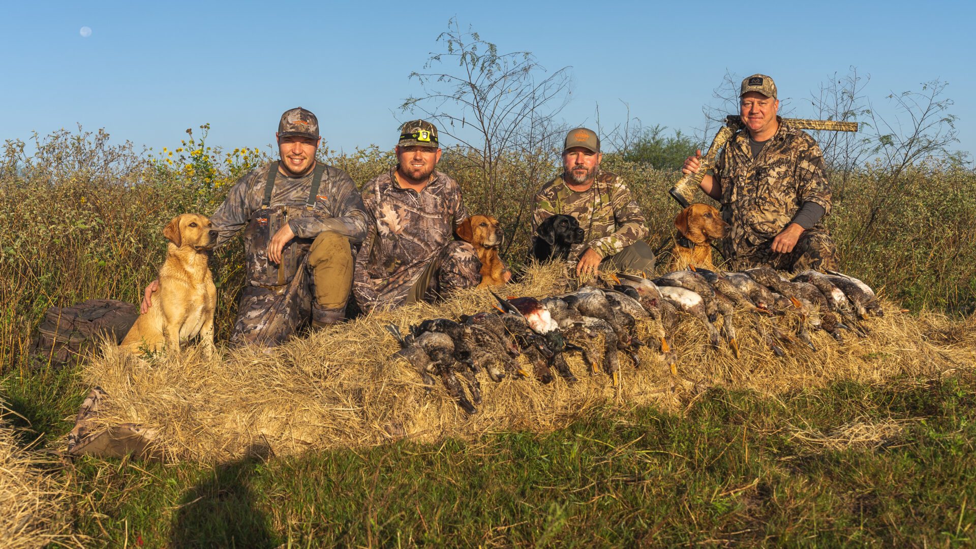 A person wearing camouflage waders and holding a shotgun stands knee-deep in water, surrounded by decoy ducks. A row of ducks can be seen in the distance. The overcast sky casts a muted light over the scene, creating a serene and focused atmosphere.