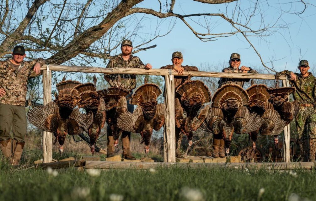 A man in camouflage clothing and a cap is holding two wild turkeys by their tails in an outdoor setting with grass, trees, and a body of water in the background. The man appears to be sitting on a rusty yellow metal beam while smiling at the camera.